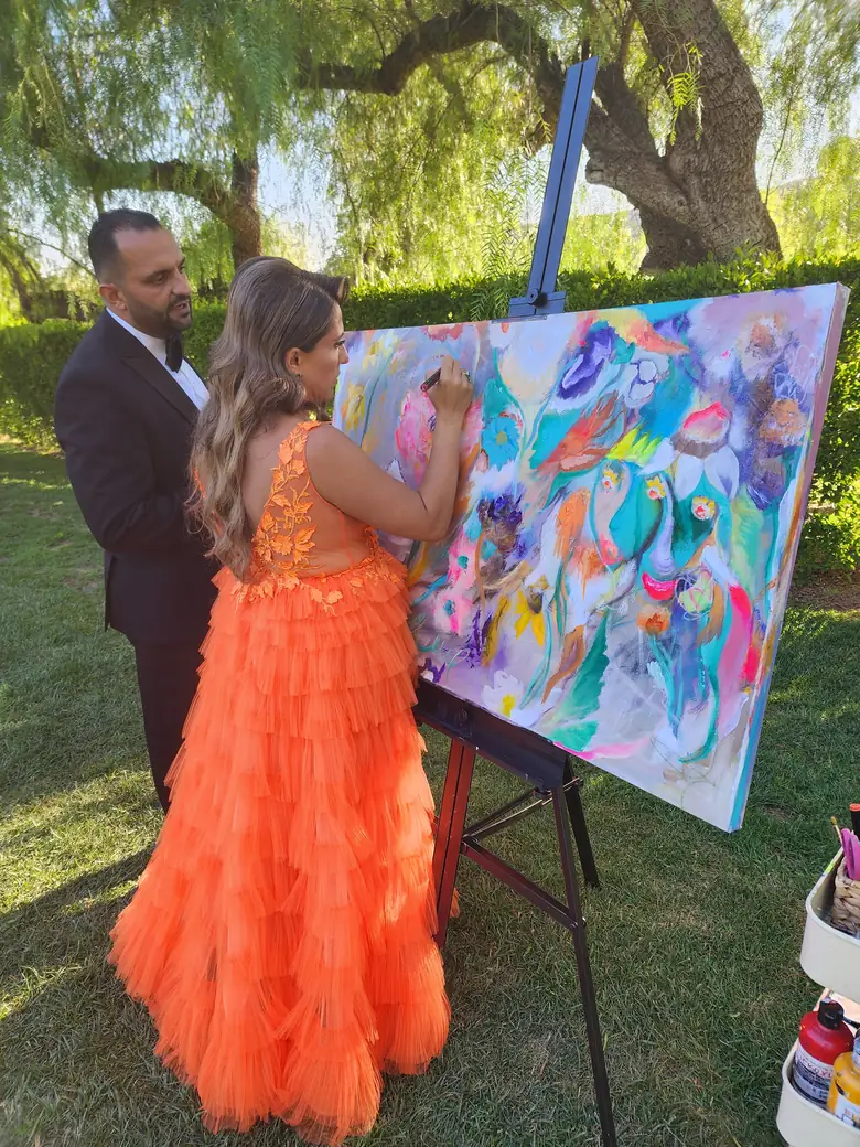 A bride in an orange gown and her groom paint together on their wedding canvas in a garden
